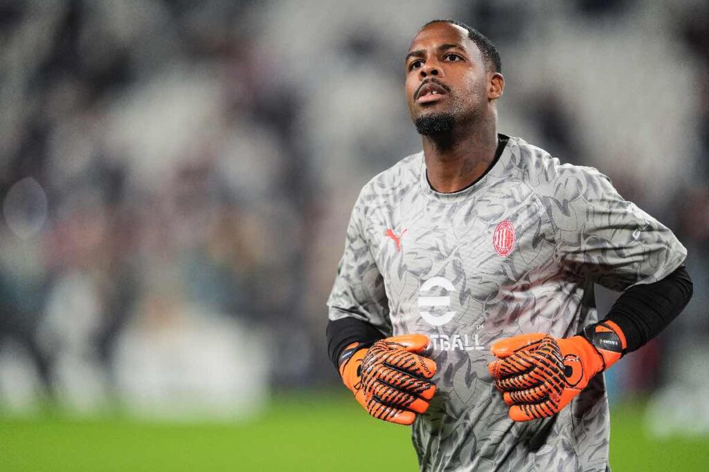 Mike Maignan, portiere del Milan, si prepara per la partita durante un riscaldamento pre-gara allo stadio. La scena mostra Maignan con il suo completo da portiere mentre guarda l'azione in lontananza, con un'espressione concentrata.