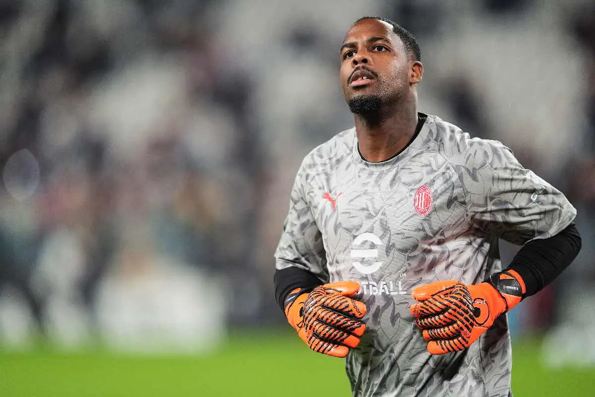 Mike Maignan, portiere del Milan, si prepara per la partita durante un riscaldamento pre-gara allo stadio. La scena mostra Maignan con il suo completo da portiere mentre guarda l'azione in lontananza, con un'espressione concentrata.