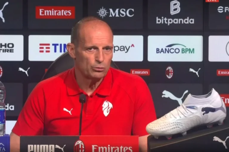 Massimiliano Allegri in conferenza stampa con microfono, indossa la polo rossa del Milan davanti ai marchi sponsor