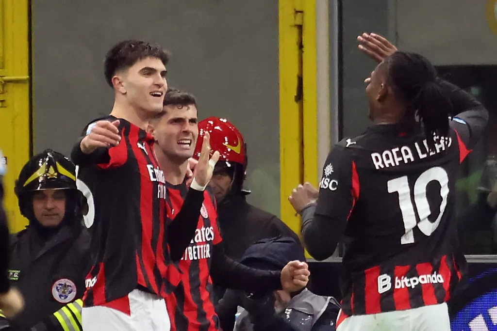 Pulisic e Leao ai box durante una partita del Milan, sorridenti e a braccia aperte verso i compagni