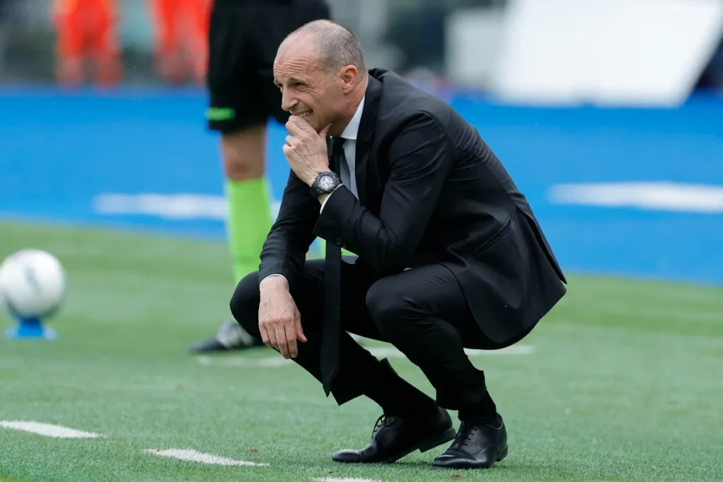 Massimiliano Allegri accovacciato in panchina durante una partita, con la mano al viso in atteggiamento pensieroso