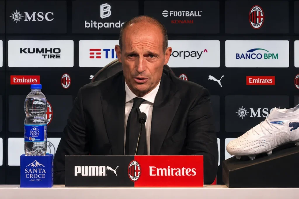 Massimiliano Allegri durante una conferenza stampa con microfono, water bottle e scarpe da calcio sullo sfondo Milan