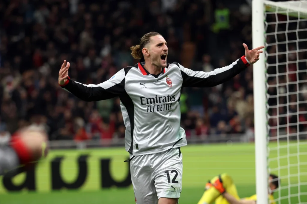 Adrien Rabiot in maglia bianca del Milan con braccia aperte durante una partita, accanto al palo della porta