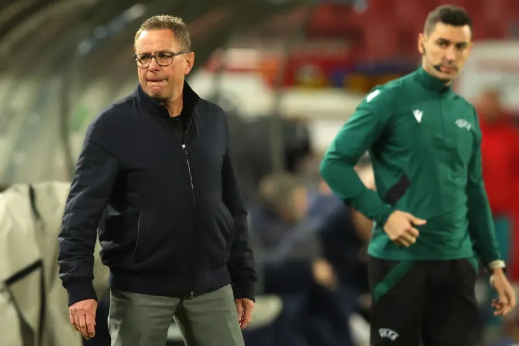 Ralf Rangnick in giacca blu scuro durante una sessione di allenamento, con un calciatore in maglia verde sullo sfondo