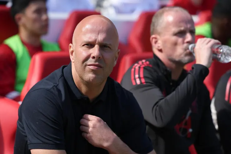Allenatore in panchina durante una partita, con staff tecnico sullo sfondo e sedie rosse dello stadio