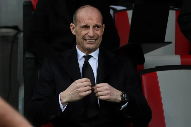 Massimiliano Allegri in panchina sorride durante una partita, indossa completo scuro con cravatta nera