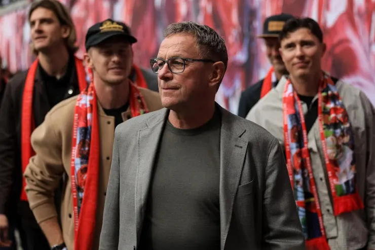 Rangnick in primo piano con occhiali e giacca grigia, circondato da tifosi rossoneri con sciarpe del Milan sullo sfondo