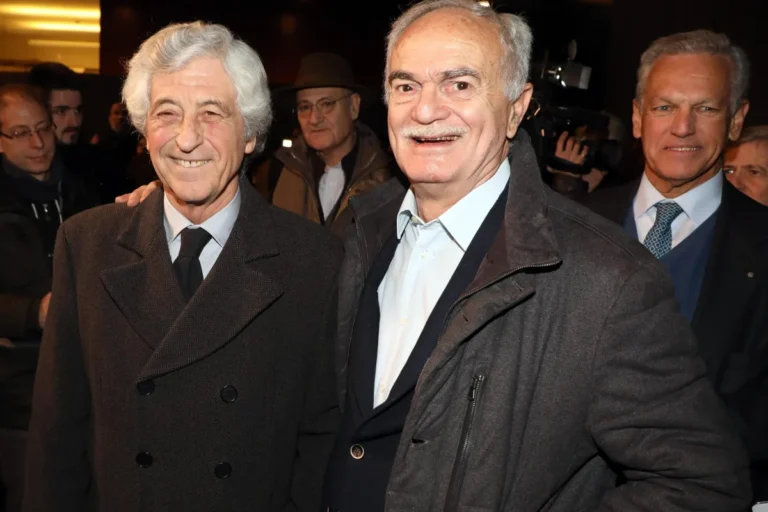 Sandro Mazzola sorride in primo piano con personalità del calcio in un evento serale, circondato da colleghi e fotografi