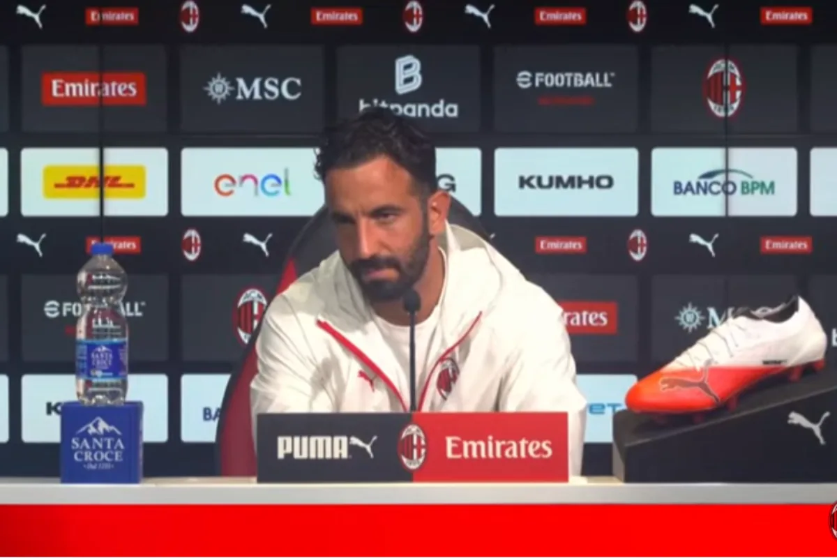 Conferenza stampa del Milan: uomo barbuto in giacca bianca al tavolo, microfono davanti e scarpa Puma sulla destra