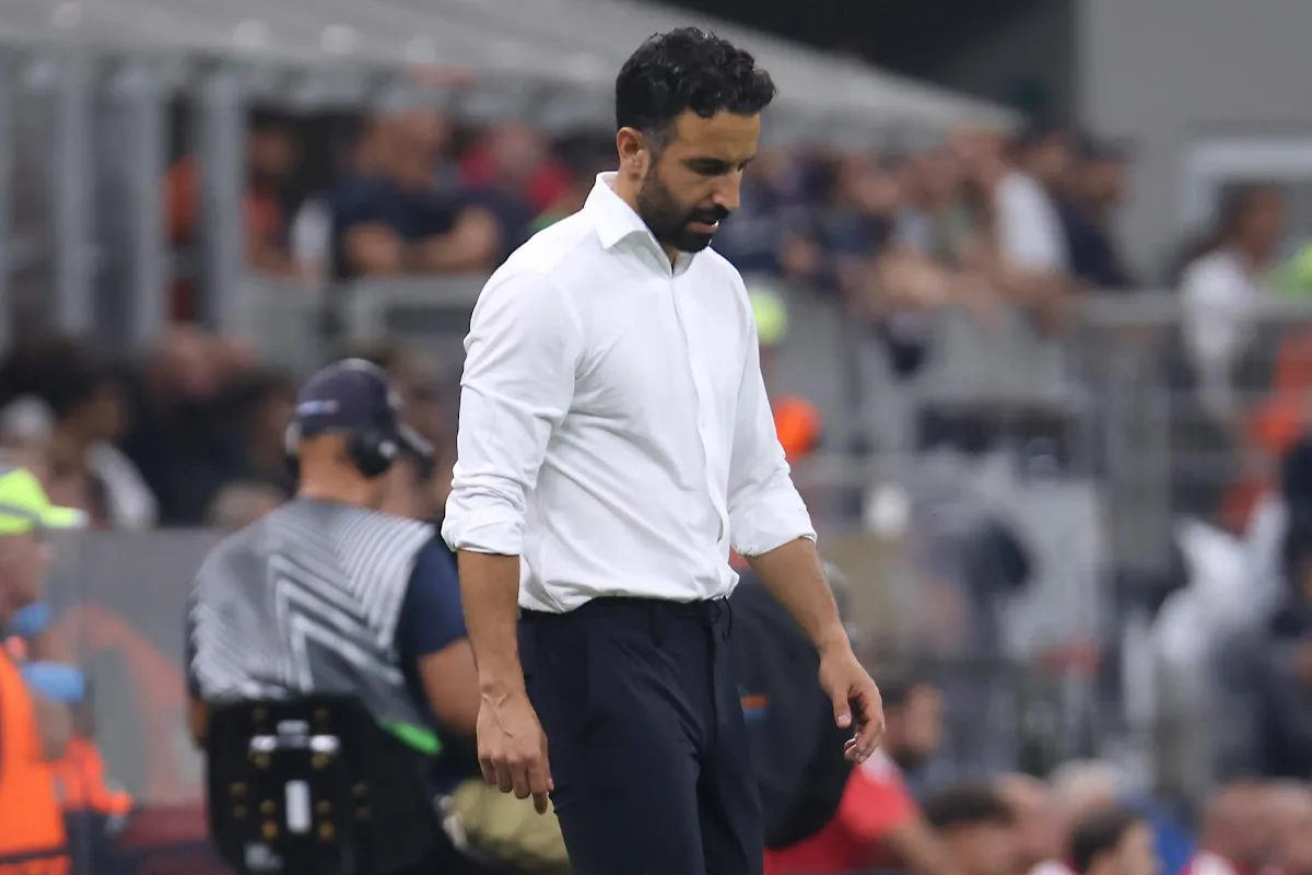 Allenatore in camicia bianca e pantaloni scuri a bordo campo, con lo sguardo abbassato durante una partita
