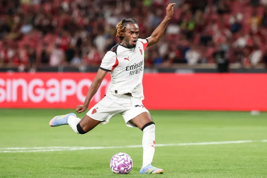 Bondo in campo con la maglia bianca del Milan mentre calcia il pallone, con sponsor Emirates e tifosi sullo sfondo