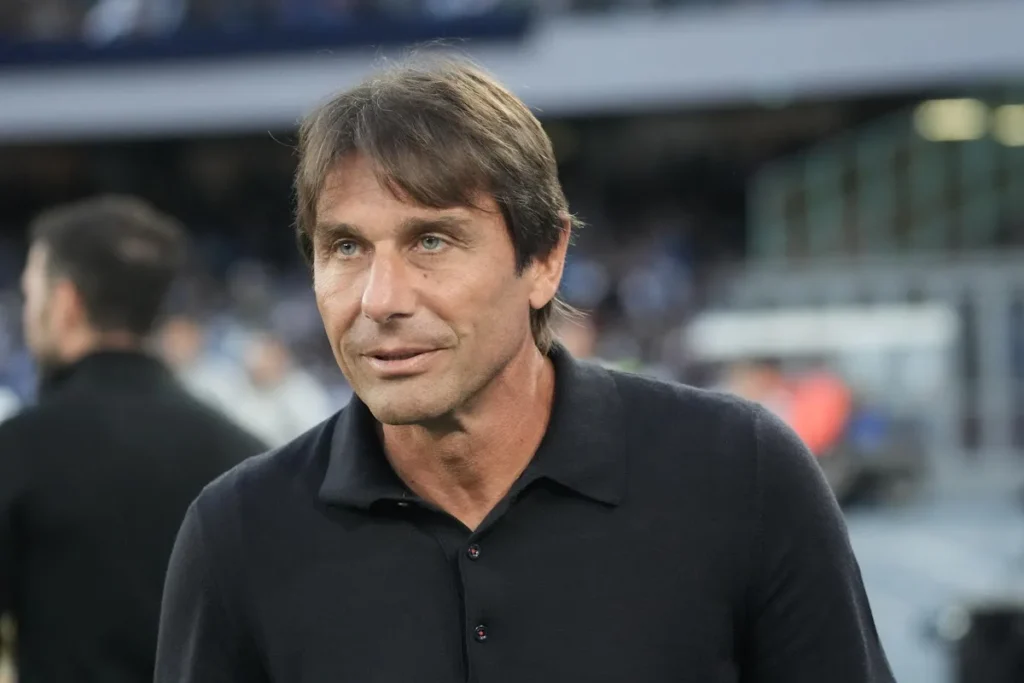 Antonio Conte in primo piano durante una partita, indossa una polo nera e osserva il campo da bordo