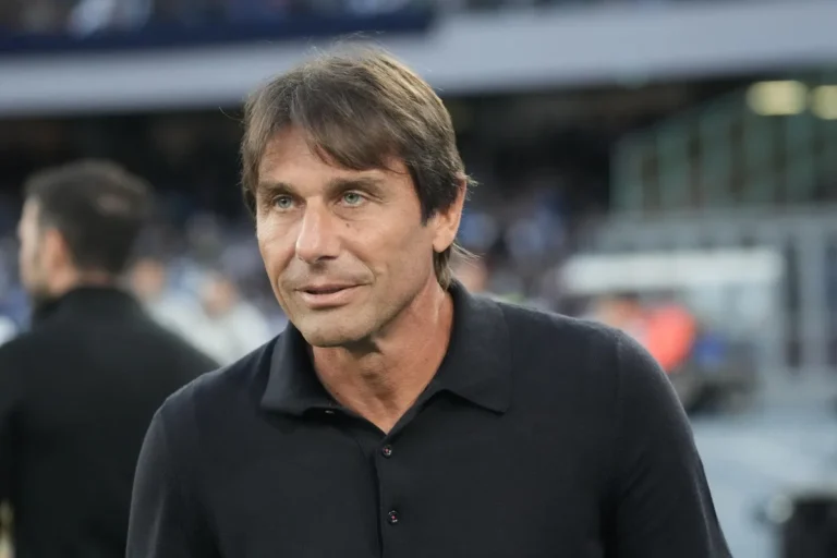 Antonio Conte in primo piano durante una partita, indossa una polo nera e osserva il campo da bordo