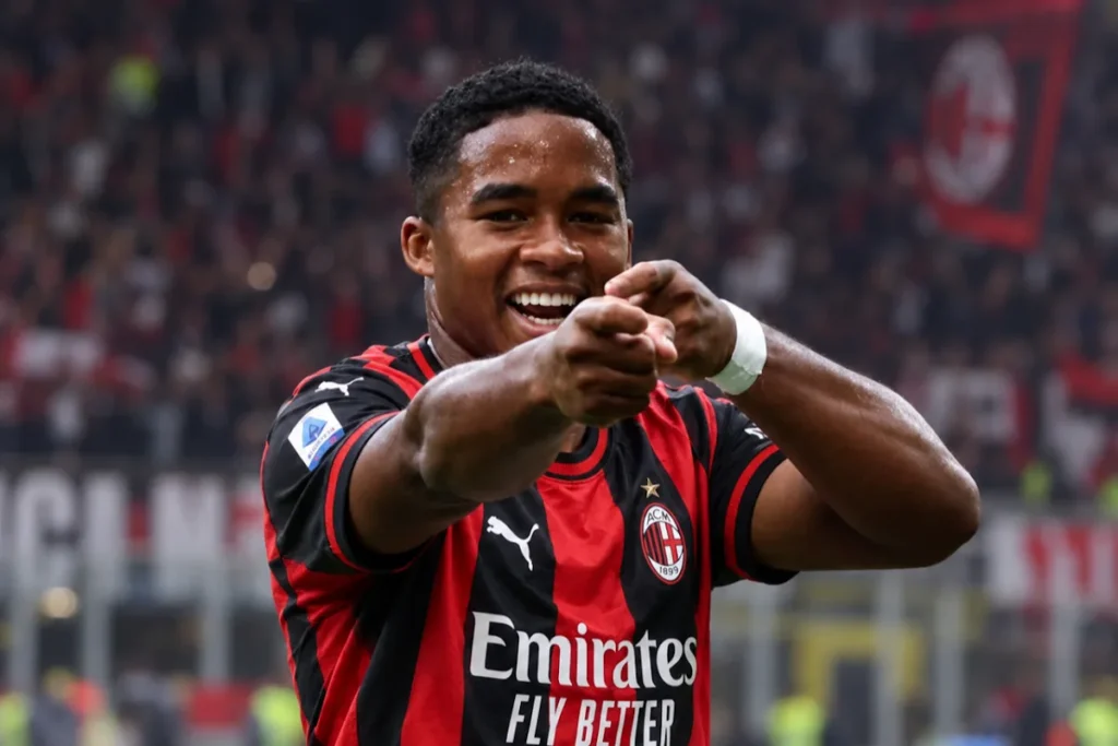 Calciatore del Milan con maglia rossonera Puma e sponsor Emirates, sorride e indica verso i tifosi