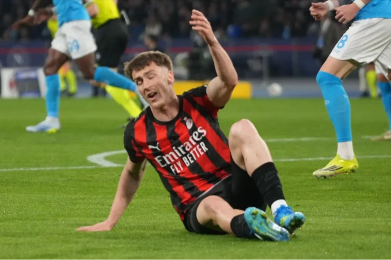 Giocatore del Milan in maglia rossonera a terra durante una partita, con avversari in blu sullo sfondo