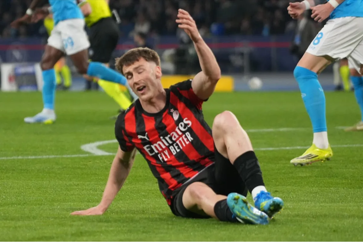 Giocatore del Milan in maglia rossonera a terra durante una partita, con avversari in blu sullo sfondo