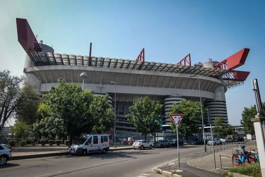 Esterno dello stadio San Siro di Milano con le caratteristiche coperture rosse in primo piano e auto in transito